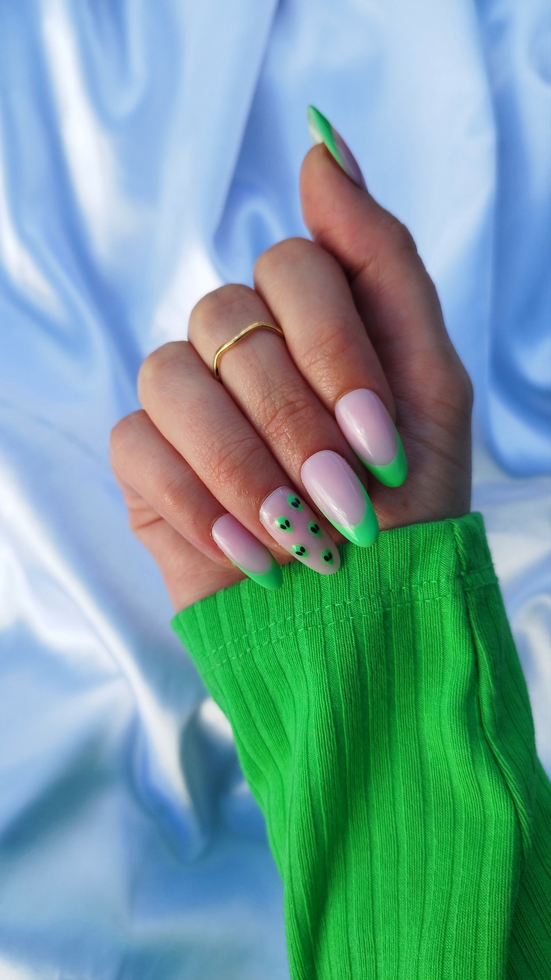 May include: Close-up of a hand with long, almond-shaped nails painted with a light pink polish. The nails have a green tip with black dots on the nail bed.