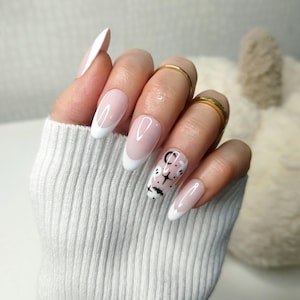 May include: A hand with long, oval-shaped nails painted with a white french manicure. The ring finger nail has a design of ghosts, stars, and moons in black.