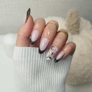 May include: Close-up of a hand with oval-shaped nails painted with a black and white design. The nails are painted with a light pink base and have a black half-moon shape at the base. The design on the nails features black and white abstract shapes and lines.