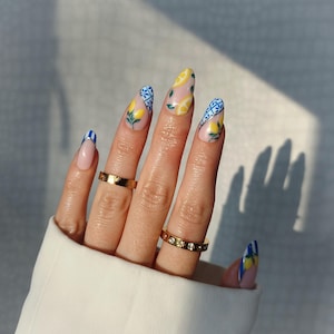 Puede incluir: Una mano con uñas largas en forma de almendra con un diseño de limón. Las uñas están pintadas con una base transparente, limones amarillos, patrones azules y blancos y una manicura francesa. Dos anillos dorados adornan los dedos.