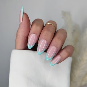 Puede incluir: Uñas artificiales ovaladas con base rosa claro y punta francesa turquesa. Se ve un anillo dorado en un dedo. Las uñas están bien cuidadas. La mano descansa sobre una superficie blanca.