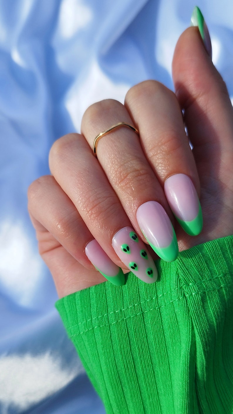 May include: Close-up of a hand with light pink oval-shaped nails with a green tip and black alien face designs.
