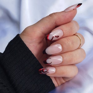 May include: Close-up of a hand with red and black flame-shaped nail art on almond-shaped nails.
