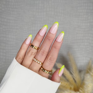 May include: Close-up of a hand with five nails painted with a light pink base and a neon yellow tip. The hand is wearing three gold rings.