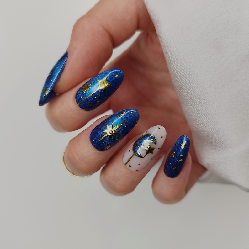 May include: A hand displaying a set of long, oval-shaped nails. The nails are painted in a deep blue with gold star and moon designs. One nail features a light pink base with a gold moon and star design.