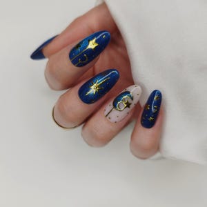 May include: Oval-shaped, dark blue nail art with gold star and crescent moon designs. One nail is light pink with gold dots and a gold moon and star. The nails have a glossy finish, creating a celestial theme.