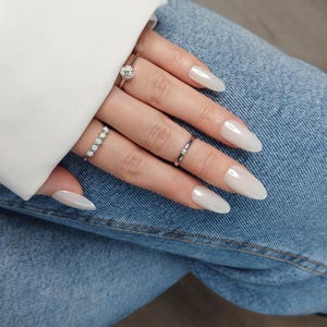 May include: Close-up of a hand with long, oval-shaped, white nails. The hand is wearing several rings, including a ring with a small, round, clear stone.