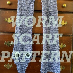 Puede incluir: Una bufanda de punto gris con un dise&ntilde;o de gusano y ojos saltones. La bufanda est&aacute; colgada en una c&oacute;moda de madera con cajones.