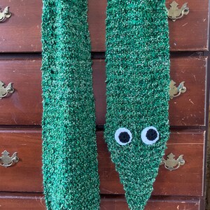 Puede incluir: Una bufanda de punto verde con un par de ojos blancos unidos al extremo. La bufanda est&aacute; colgada de una c&oacute;moda de madera.