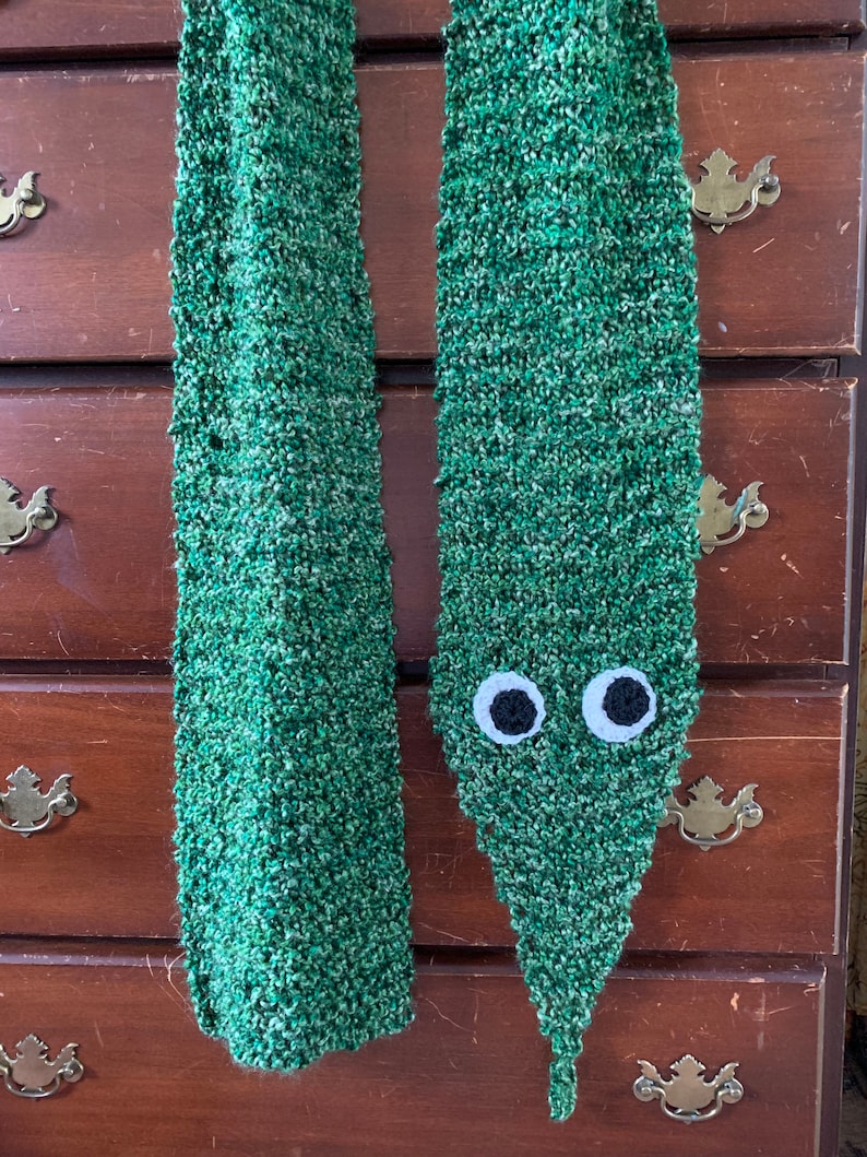 Puede incluir: Una bufanda de punto verde con un par de ojos blancos unidos al extremo. La bufanda est&aacute; colgada de una c&oacute;moda de madera.