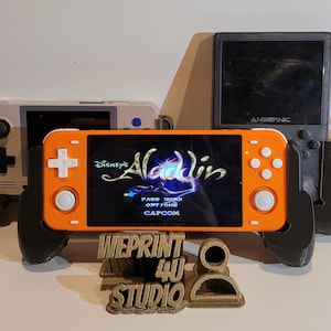 May include: A collection of handheld video game consoles, including a bright orange console with a screen displaying the game "Disney's Aladdin". The consoles are displayed on a black stand with a gold 3D printed logo that reads "WEPRINT 4U STUDIO".