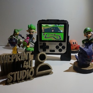 May include: A black handheld video game console with a green and brown racing game displayed on the screen. The console is on a black stand. There are several small green, brown, and purple figurines in the background. The text "WEPRINT 4U STUDIO" is printed on a brown object in the foreground.