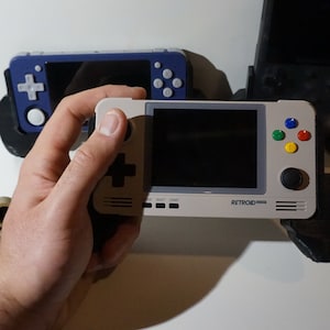 May include: A white and gray handheld gaming console with a black screen, a directional pad, four colored buttons, and a circular button. The console has the text "RETROID POCKET" printed on it.