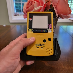May include: A yellow Game Boy Color handheld video game console with a Pokemon game cartridge inserted. The console has a black screen and a white sticker with the words "GAME BOY COLOR" and "Nintendo" on it. The game cartridge is yellow with a blue Pokemon logo and the words "Pokemon" and "Game Freak" on it.
