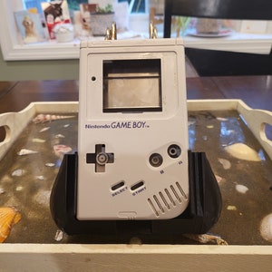 Könnte beinhalten: Eine weiße Nintendo Game Boy Handheld-Spielkonsole mit einem schwarzen und grauen Ständer. Die Konsole hat einen grauen Bildschirm, einen schwarzen Steuerkreuz und schwarze Knöpfe mit den Beschriftungen "Select", "Start", "A" und "B".