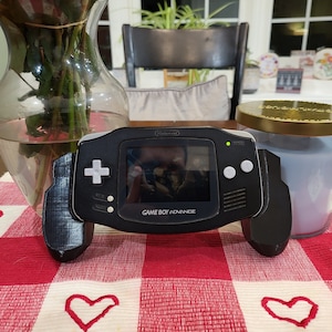 May include: A black Game Boy Advance handheld gaming console with a screen displaying a video game. The console has a white button on the right side and a white directional pad on the left side. The console is sitting on a red and white checkered table runner.