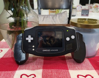 May include: A black Game Boy Advance handheld gaming console with a screen displaying a video game. The console has a white button on the right side and a white directional pad on the left side. The console is sitting on a red and white checkered table runner.