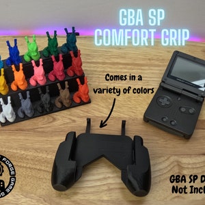May include: A black 3D printed Game Boy Advance SP comfort grip. The grip is designed to make the handheld console more comfortable to hold. The grip is shown on a wooden surface with a Game Boy Advance SP console and a variety of coloured 3D printed figures. The text "GBA SP COMFORT GRIP" is shown in the upper right corner of the image. The text "Comes in a variety of colours" is shown in the middle of the image. The text "GBA SP Device Not Included" is shown in the lower right corner of the image.