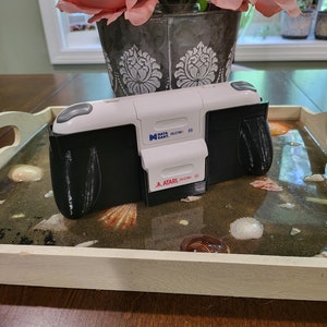 May include: A white and black Atari video game console with a Data East cartridge inserted. The console is sitting on a tray with a seashell design.