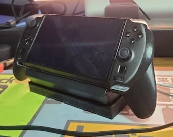May include: A black handheld gaming console with a screen and buttons. The console is sitting on a black stand.