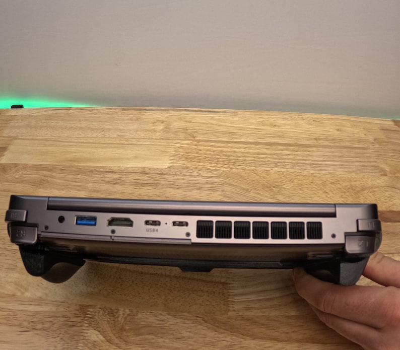 May include: A dark gray laptop with a black rubberized grip on each side. The laptop has a USB port, HDMI port, and a power port on the side. The laptop is sitting on a wooden surface.