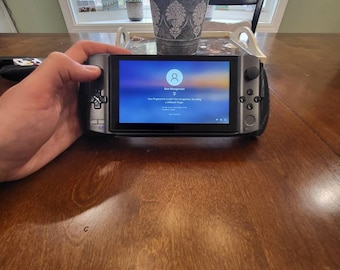 May include: A black handheld gaming device with a screen displaying a login prompt. The screen shows a user icon and the text "Sign in to Windows". The device has a gray trim and buttons on the sides.