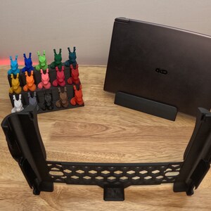 May include: A black laptop stand with a laptop on top of it. The laptop is closed and has a black lid. There are 20 small, colorful figurines on a shelf in the background. The stand is made of black plastic and has a honeycomb design.