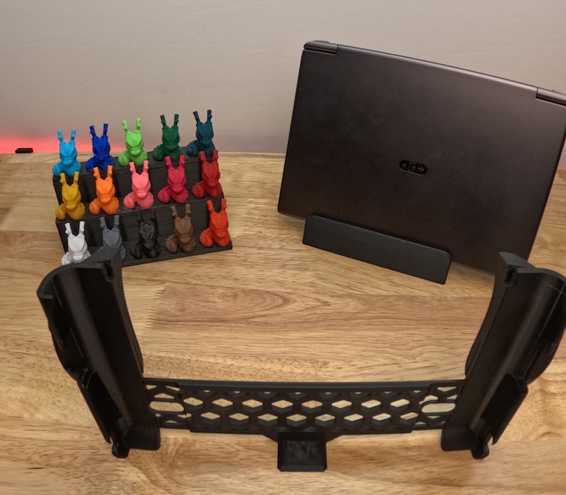 May include: A black laptop stand with a laptop on top of it. The laptop is closed and has a black lid. There are 20 small, colorful figurines on a shelf in the background. The stand is made of black plastic and has a honeycomb design.