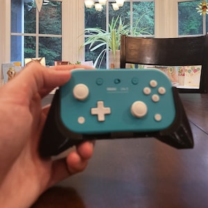 May include: A teal blue Nintendo Switch Joy-Con controller with white buttons and a black grip. The controller is being held in a hand.