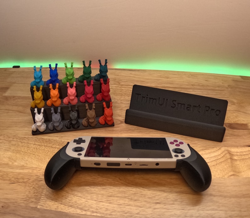 K&ouml;nnte beinhalten: Ein schwarzer St&auml;nder mit dem Text "TrimUI Smart Pro" h&auml;lt eine wei&szlig;e und schwarze Handheld-Spielkonsole mit schwarzen Griffen. Eine bunte Auswahl von 20 kleinen, 3D-gedruckten Figuren steht auf einer Holzoberfl&auml;che.
