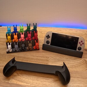 K&ouml;nnte beinhalten: Ein schwarzer St&auml;nder mit einer grauen Handheld-Spielkonsole, einem schwarzen Controller und einem mehrfarbigen 3D-gedruckten Displayst&auml;nder mit kleinen Figuren.