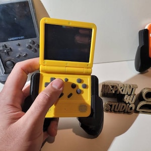 May include: A yellow handheld gaming console with a black controller. The console has a screen, buttons, and a directional pad. The console is being held by a hand.
