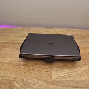 May include: A closed, dark gray laptop computer with a black plastic laptop stand. The laptop is sitting on a light wood surface.