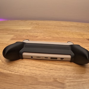 K&ouml;nnte beinhalten: Eine schwarz-wei&szlig;e Handheld-Spielkonsole mit einer schwarzen Schutzh&uuml;lle. Die Konsole hat eine wei&szlig;e Frontplatte mit Anschl&uuml;ssen und Kn&ouml;pfen.