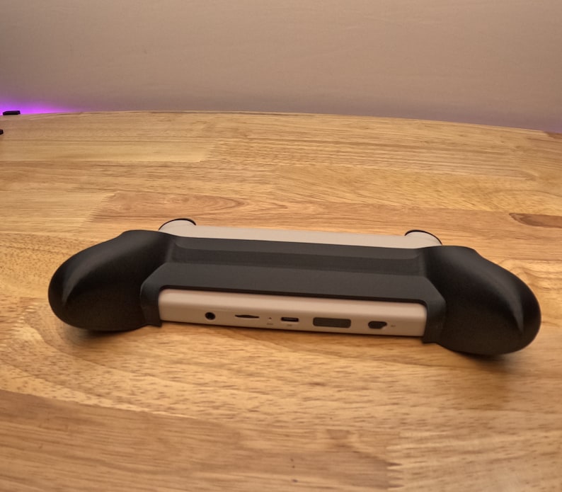 K&ouml;nnte beinhalten: Eine schwarz-wei&szlig;e Handheld-Spielkonsole mit einer schwarzen Schutzh&uuml;lle. Die Konsole hat eine wei&szlig;e Frontplatte mit Anschl&uuml;ssen und Kn&ouml;pfen.