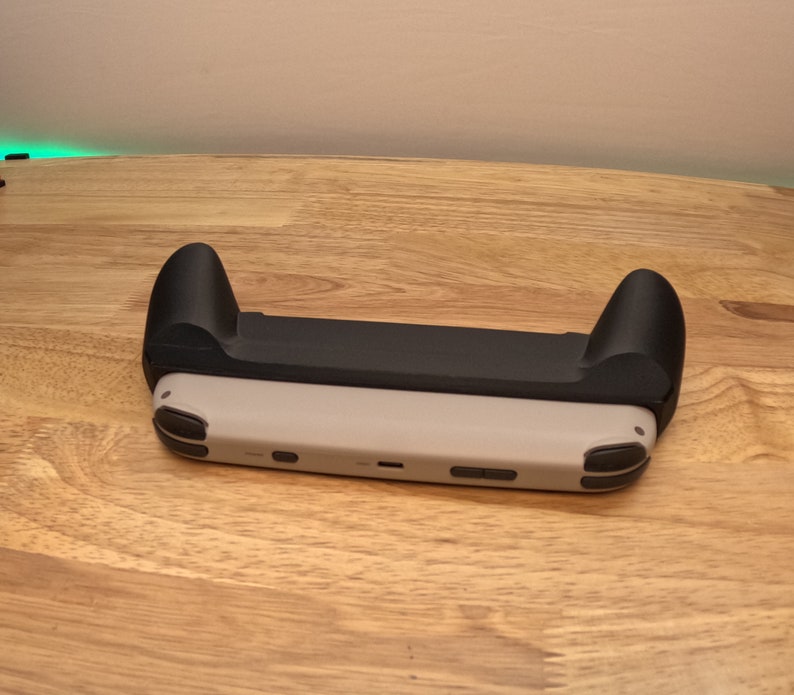 K&ouml;nnte beinhalten: Eine schwarze und graue Nintendo Switch-Konsole mit einem schwarzen Griffaufsatz. Die Konsole liegt auf einer Holzoberfl&auml;che.