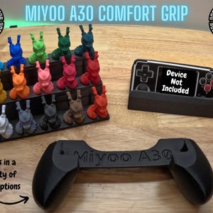 May include: A black Miyoo A30 handheld gaming console comfort grip with the text "Miyoo A30" printed on the top. The grip is shown on a wooden surface. A display stand for a handheld gaming console is also shown in the image. The stand is black and has the text "Device Not Included" printed on it. A variety of colored 3D printed figures are shown in the background.