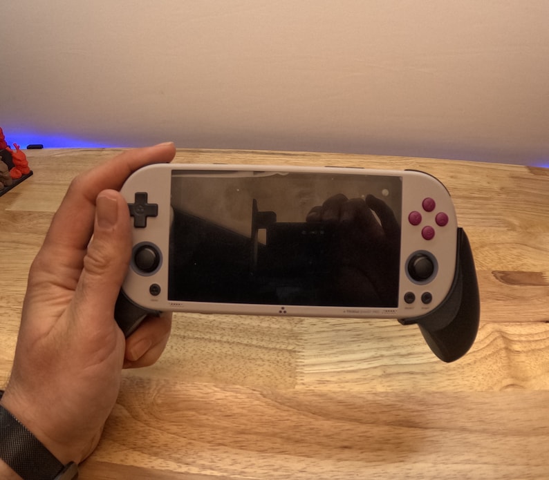 K&ouml;nnte beinhalten: Eine wei&szlig;e und graue Handheld-Spielkonsole mit schwarzem Geh&auml;use. Die Konsole hat einen schwarzen Bildschirm, einen violetten Knopf und einen schwarzen Joystick.