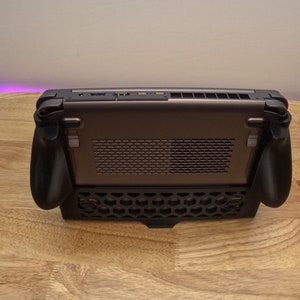 May include: A black handheld gaming device with a honeycomb pattern on the bottom and two black controllers on the sides. The device has a silver back with a black grill.