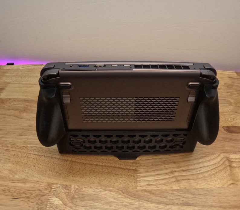 May include: A black handheld gaming device with a honeycomb pattern on the bottom and two black controllers on the sides. The device has a silver back with a black grill.
