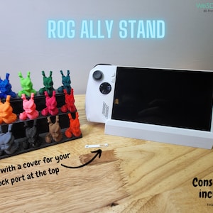 Könnte beinhalten: Ein weißer ROG Ally Handheld-Gaming-Ständer mit schwarzem Sockel. Der Ständer hat eine kleine Abdeckung für den Docking-Port oben. Die Konsole ist nicht im Lieferumfang enthalten. Der Text "ROG ALLY STAND" ist in Blau auf weißem Hintergrund gedruckt. Der Text "Konsole nicht im Lieferumfang enthalten" ist in Schwarz auf weißem Hintergrund gedruckt.