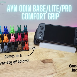 May include: A black handheld gaming console with a comfort grip, resting on a white stand. A display of colorful 3D-printed accessories in various colors is also shown. Text reads "AYN ODIN BASE/LITE/PRO COMFORT GRIP" and "Comes in a variety of colors!"