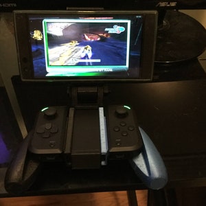May include: A black phone with a green screen is mounted to a black stand with a black controller on either side. The phone is displaying a video game.