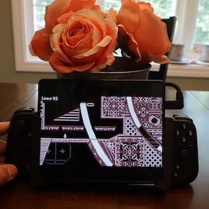 May include: A black handheld gaming device with a screen displaying a video game. The game is a side-scrolling platformer with a pixelated art style. The screen shows a level with a maze-like design and the text "Level 92".