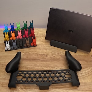 May include: A set of 18 colorful 3D printed figurines arranged on a shelf, a black laptop stand, and a black plastic accessory for a gaming console.