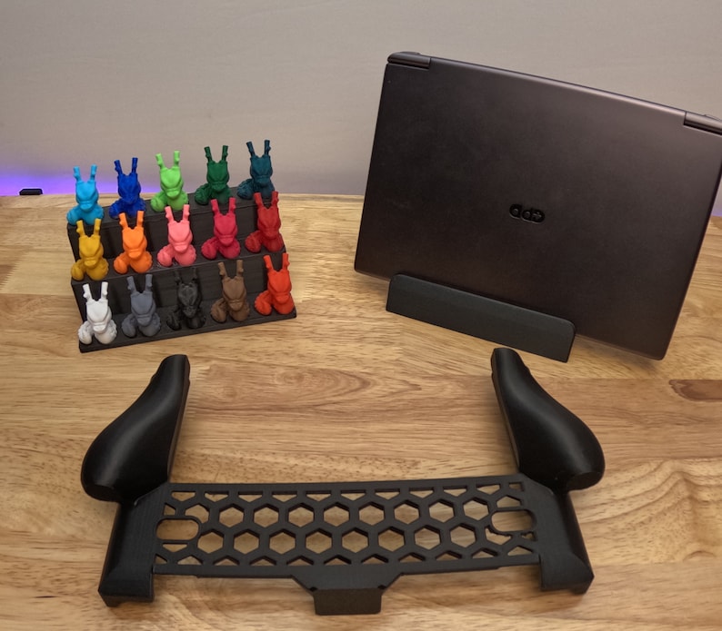 May include: A set of 18 colorful 3D printed figurines arranged on a shelf, a black laptop stand, and a black plastic accessory for a gaming console.