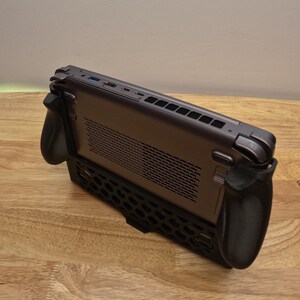 May include: A dark gray handheld gaming console with black detachable controllers. The console has a honeycomb-patterned back panel and a ventilation grill on the top.