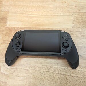 Puede incluir: Una consola de juegos port&aacute;til negra con una pantalla y botones. La consola tiene un agarre texturizado en ambos lados.