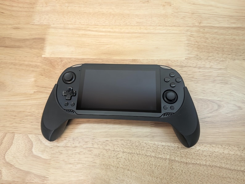 Puede incluir: Una consola de juegos port&aacute;til negra con una pantalla y botones. La consola tiene un agarre texturizado en ambos lados.