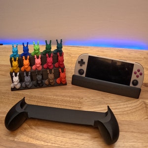 K&ouml;nnte beinhalten: Ein schwarzer St&auml;nder mit einer grauen Handheld-Spielkonsole, einem schwarzen Controller und einem mehrfarbigen 3D-gedruckten Displayst&auml;nder mit kleinen Figuren.